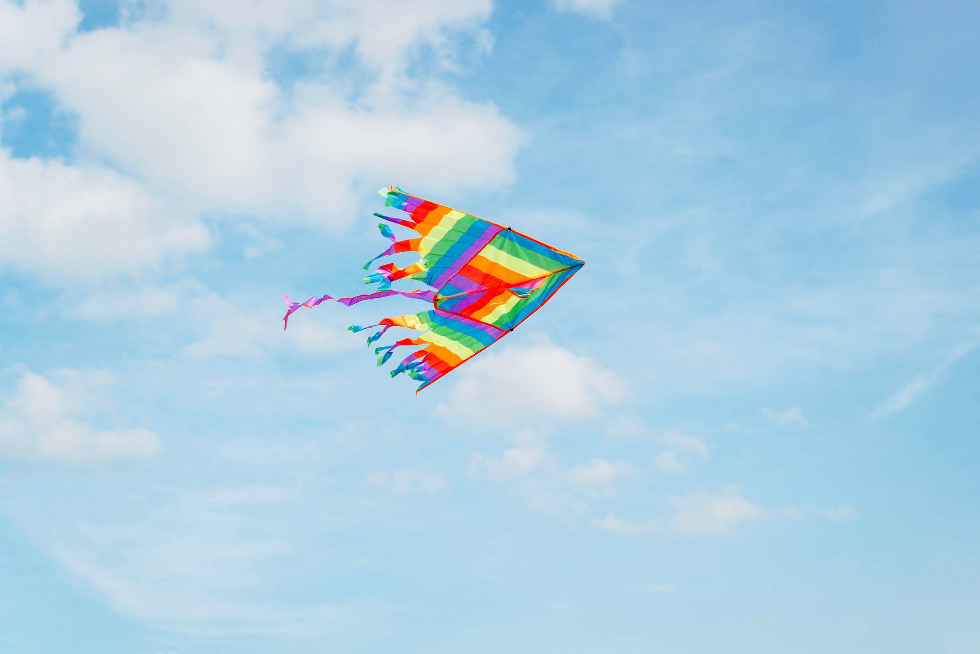Rainbow kite high in blue cloudy sky, sunny day outdoors. Color kite flying, fun and games in nature
