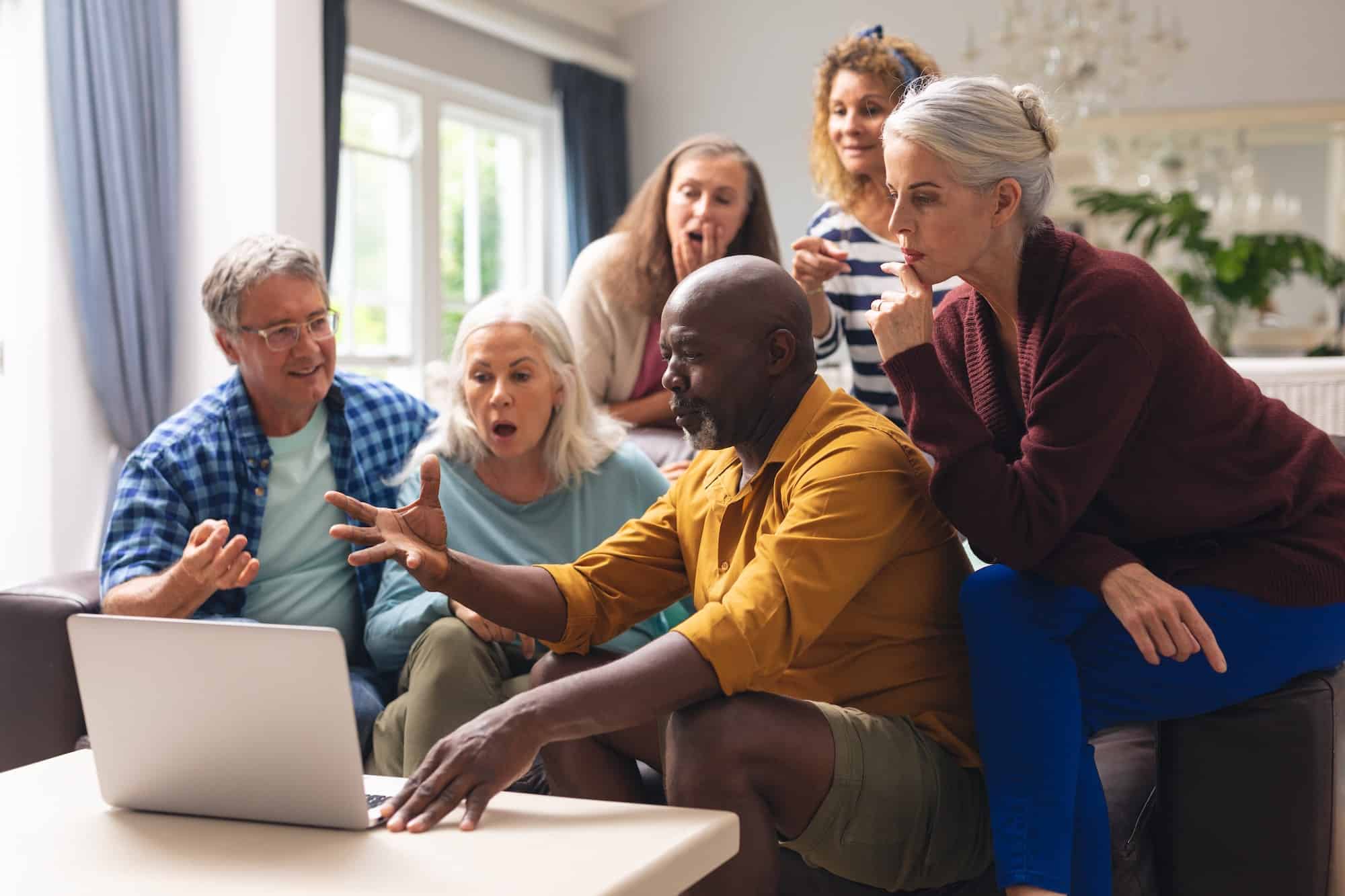 Senior african american man showing laptop to caucasian friends during social gathering at home
